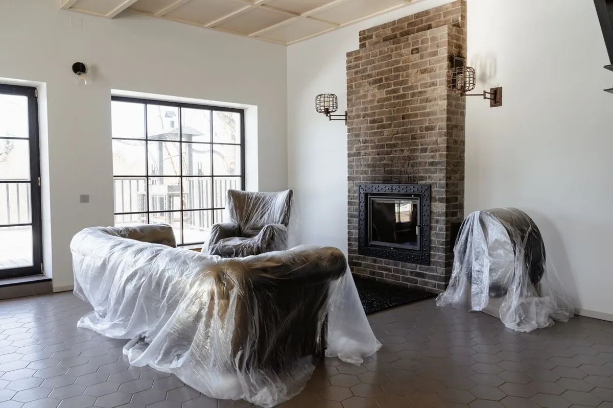 Salón con los sillones cubiertos con plásticos protectores frente a una chimenea de ladrillo